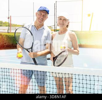Happy elderly couple with tennis racket in hand standing by net. Stock Photo