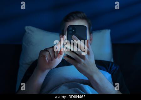 Close up of young man, alone in the dark bedroom, holds his smartphone close to face while scrolling social media or texting late at night. Screen dependence, excessive consumption of content Stock Photo