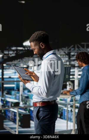 African american AI technician analyzes infrastructure performance outputs on tablet while he stands on industrial platform. Server room employee finetuning mainframes configurations. Stock Photo