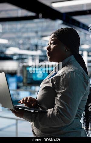 Woman in server farm using laptop, automating system diagnostics using machine learning predictions. Female technician in data center using notebook to configure gear for AI tasks Stock Photo