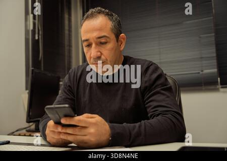 Middle-aged man sitting at a desk, intently using his smartphone screen Stock Photo