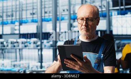 Elderly senior engineer in smart solar panels manufacturing factory uses software on tablet to maximize power generation. Experienced professional overseeing industry 4.0 photovoltaics plant, camera A Stock Photo