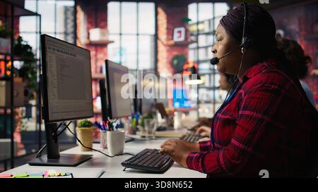 IT support specialist at PC in office helping customers during call, troubleshooting errors. Tech support agent at desk looking at computer monitor, fixing technical issues for clients, camera B Stock Photo