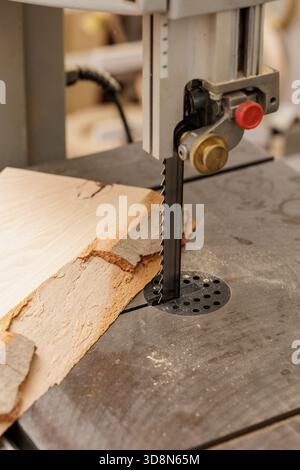 Vertical bandsaw blade cutting raw wood plank with bark edge on metal table in dusty woodworking shop symbol of timber processing and safety Stock Photo