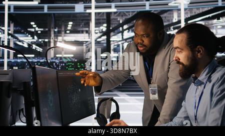 Server farm manager overseeing engineer examining neural network algorithms. Data center team leader and worker reviewing performance dashboards of artificial intelligence infrastructure, camera A Stock Photo