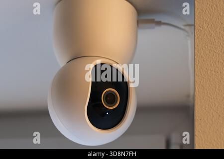 Modern white smart home security camera mounted on ceiling for indoor video surveillance and safety monitoring. Stock Photo
