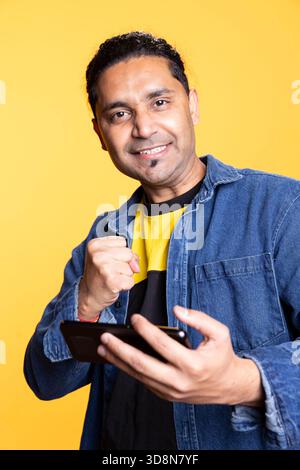 Satisfied pleased guy winning video games and feeling thrilled with the competition, enjoying interactive online multiplayer game for entertainment. Young indian man on studio background. Stock Photo