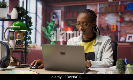 Teleworker analyzing data on laptop at desk and looking over paperwork, compiling analytical reports. Remote employee checking documents, using notebook to upload company information online, camera B Stock Photo