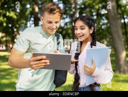 Two Young Students Share Excitement Over a Tablet in a Sunny Park While Holding Notebooks Stock Photo