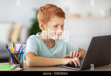 Joyful Ginger Boy Focused on Homework While Using Laptop at Modern Desk Stock Photo