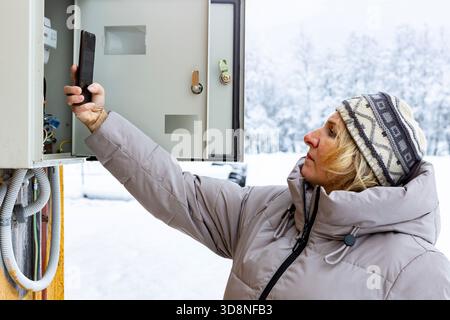 Taking meter readings homeowner in winter clothes holds gadget pointing it at power meter in service panel on outside wall of countryside home. Stock Photo
