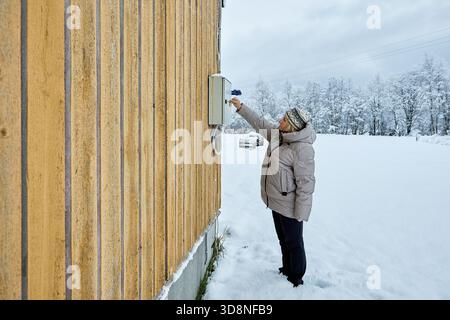 Meter data capture local resident in winter coat holds mobile device aiming it at electricity meter inside main panel on exterior wall of house. Stock Photo