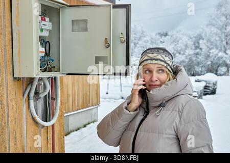 Standing near open outdoor switchboard with circuit breakers and electricity meter woman blonde middle aged in warm winter clothes uses mobile phone t Stock Photo