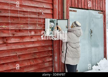 Monitoring exterior meter homeowner in winter clothing uses smartphone opposite snowy wall of countryside structure during cold season. Stock Photo