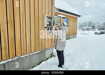 Meter reading capture via photo woman in winter clothes holds communicator photographing power meter inside switchboard on exterior wall of countrysid Stock Photo