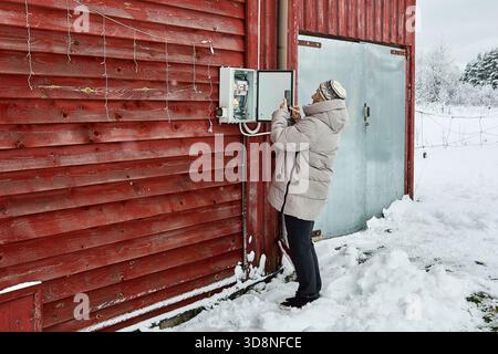 Recording meter data homeowner in winter clothing uses smartphone near snowy wall of rural building during cold season. Stock Photo