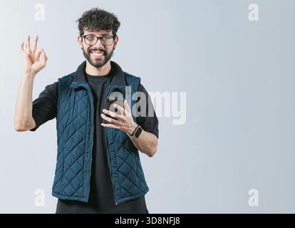 Attractive man in glasses holding phone gesturing OK. Smiling handsome man holding phone and gesturing OK, isolated Stock Photo