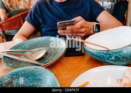 a man holds his smartphone with both hands at a restaurant table while the device stays connected to a power bank, surrounded by empty bowls and cutle Stock Photo