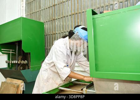 Woman wearing a lab coat and face shield operating green industrial machinery in a factory for manufacturing and production Stock Photo