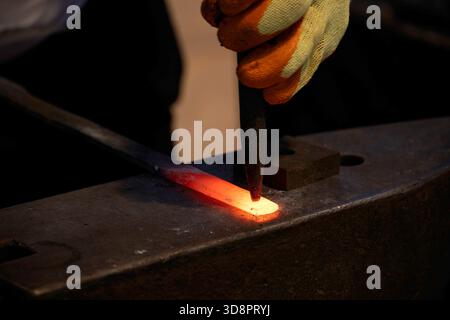 Doha, Qatar - December 01, 2025: Close up of a blacksmith shaping a glowing hot metal piece on an anvil, using a chisel while wearing protective glove Stock Photo