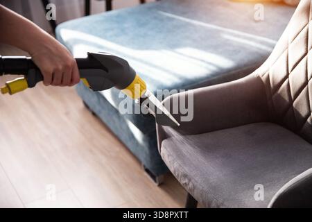 A person uses a steam cleaner to remove dirt from a gray fabric chair in a cozy living room. Sunlight streams through the window, creating a warm atmo Stock Photo