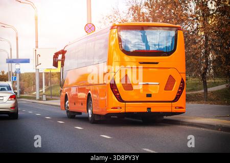 Back of large orange passenger bus stopped on roadside in urban setting with autumn trees. Sunlight creates lens flare over streetlights and another v Stock Photo