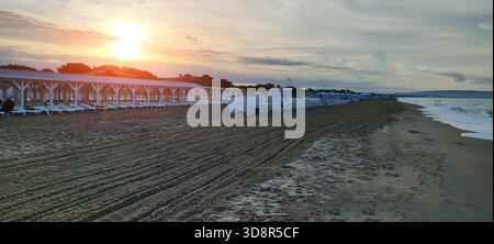 Wide view of beach at early morning or late afternoon. Bright sun on horizon casts warm glow. Long line of empty white beach chairs under white wooden Stock Photo