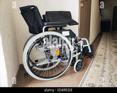 Modern manual wheelchair indoors, side view. Black fabric seat, backrest, armrests, silver frame. Large rear wheels, white tires, small front caster w Stock Photo