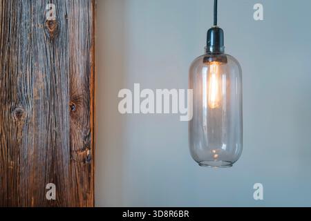 Close-up of a modern vintage-style glass pendant light fixture with a glowing filament bulb, next to an old wooden beam Stock Photo