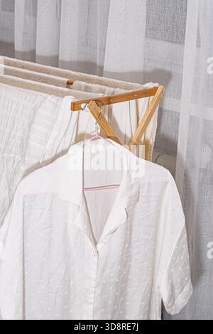 White shirt on hanger drying on bamboo laundry rack with fresh towels near window, symbolizing clean clothes, eco home care and simple lifestyle. Stock Photo