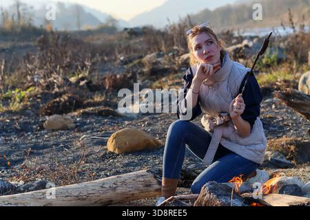Introspective moment: woman in jeans touches lips, sausage skewer rests on ground near campfire in autumn Stock Photo