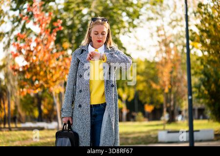 Worried businesswoman in autumn coat checking the time on her wristwatch while while walking and carrying her laptop bag through the city park. Stock Photo