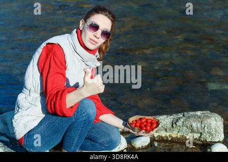 Stylish young lady in vest and sunglasses poses thumbs up next to paper container of red cherry tomatoes by flowing stream. Stock Photo
