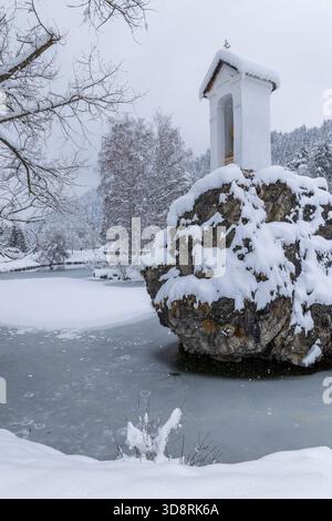 Small white chapel on a snow covered rock stands beside a frozen pond in Liptovsky Hradok, Slovakia Stock Photo