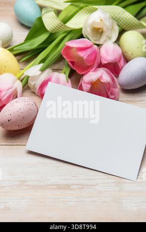 Easter greeting card is surrounded by colorful eggs and pink tulips on a white wooden table Stock Photo