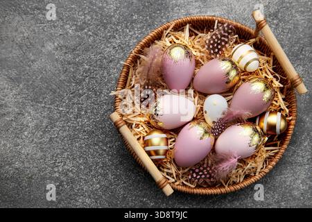 Golden and pink colored eggs and feathers in wicker plate on a dark concrete background Stock Photo