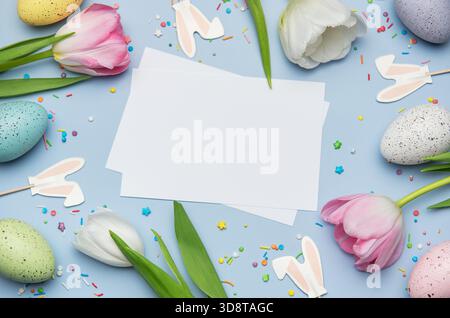 Easter greeting card surrounded by colorful eggs, tulips and bunny ears on pastel blue background with sprinkles Stock Photo