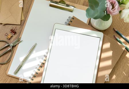 A bright workspace features a blank tablet, an open notebook, stationery tools, and fresh flowers arranged on a wooden table, inviting creativity and planning. Stock Photo