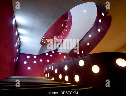 View of a modern staircase curves upwards, its deep red walls punctuated by glowing, circular lights, creating a mesmerizing visual rhythm, Suginami C Stock Photo