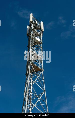 A tall telecommunications tower constructed of silver metal rises high against a clear blue sky with sparse, wispy white clouds on a bright, sunny day Stock Photo