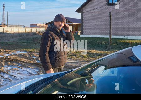 Older man talking cell phone, standing front car broken down. Calling roadside assistance driver frustrated. Emergency situation in cold winter day ag Stock Photo