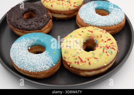 Donuts with bright icing and sprinkles on black plate. Sweet pastry dessert on white background. Stock Photo