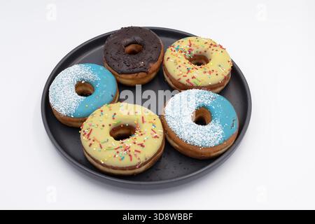 Donuts with bright icing and sprinkles on black plate. Sweet pastry dessert on white background. Stock Photo