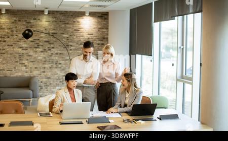Four professionals engage in a lively discussion in a modern office. Two are seated at a table with laptops, while two others stand nearby, sharing id Stock Photo