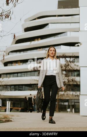 A business professional walks with purpose outside a contemporary building. She is dressed in formal attire and carrying a handbag. It is a clear day, Stock Photo