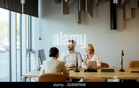 Three professionals are engaged in a meeting at a contemporary office space. They are discussing project details using digital devices and notes while Stock Photo