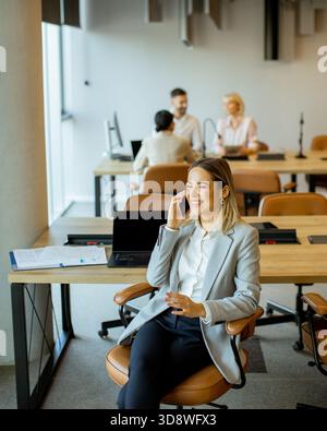 A woman in a suit smiles while talking on the phone in a bright office. Behind her, colleagues work together at tables, discussing projects and sharin Stock Photo