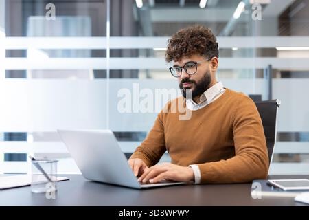 Young diverse businessman with glasses and beard working intently on a laptop at a contemporary desk in a bright, modern office, focusing on his tasks and demonstrating productivity Stock Photo