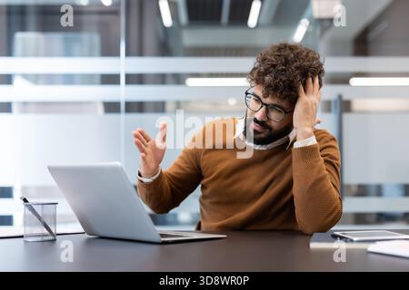 Frustrated office worker clutching his head at a laptop, showing stress, burnout and overwhelm from deadlines and technical problems in a modern corporate workspace Stock Photo