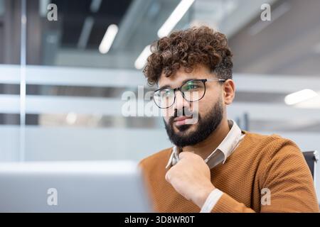 Young businessman with glasses and curly hair stares thoughtfully out the window in a modern office, focused on planning strategy and solving business challenges on his laptop Stock Photo